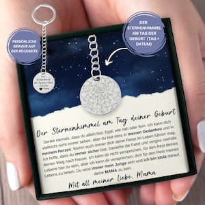 May include: A silver keychain with a round charm featuring a star constellation design. The charm is attached to a silver chain and a silver key ring. The keychain is in a black box with a white background and text that reads "Der Sternenhimmel am Tag deiner Geburt" and "Mit all meiner Liebe, Mama".