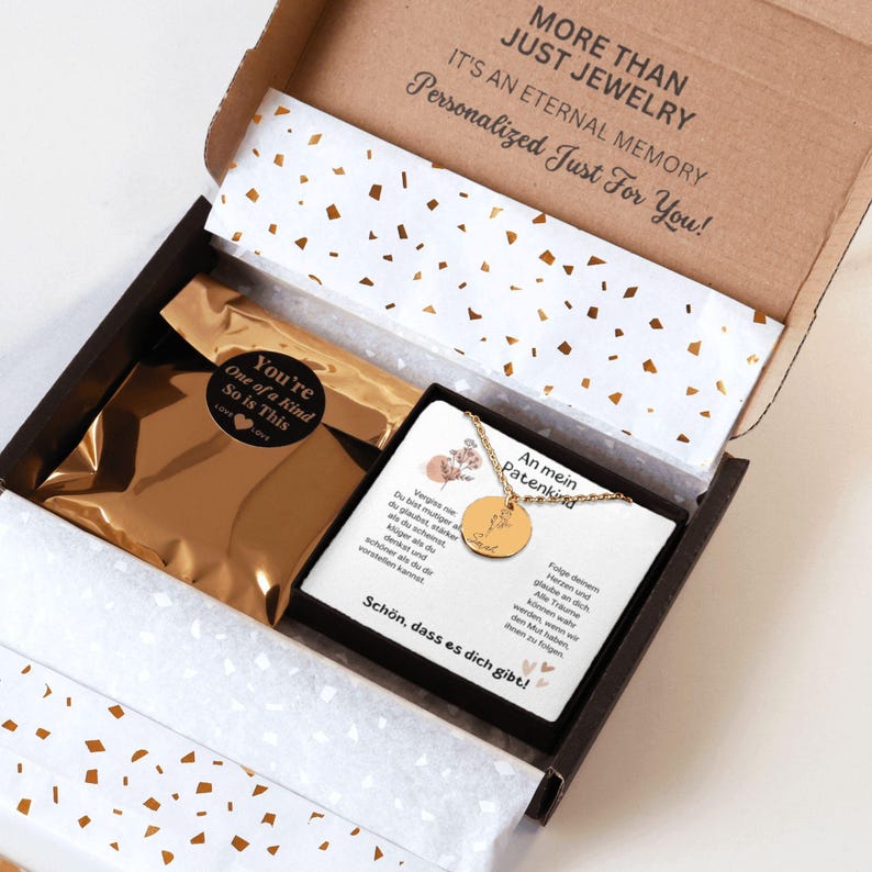 a gift box with a gold-colored interior, containing a necklace and a card. The card appears to be a personalized message or note, and the box is decorated with gold confetti.