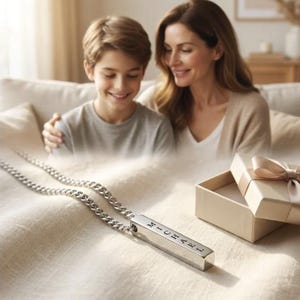 A mother and son are sitting together on a couch, smiling and looking at a necklace with a pendant that has the name "Michael" engraved on it.