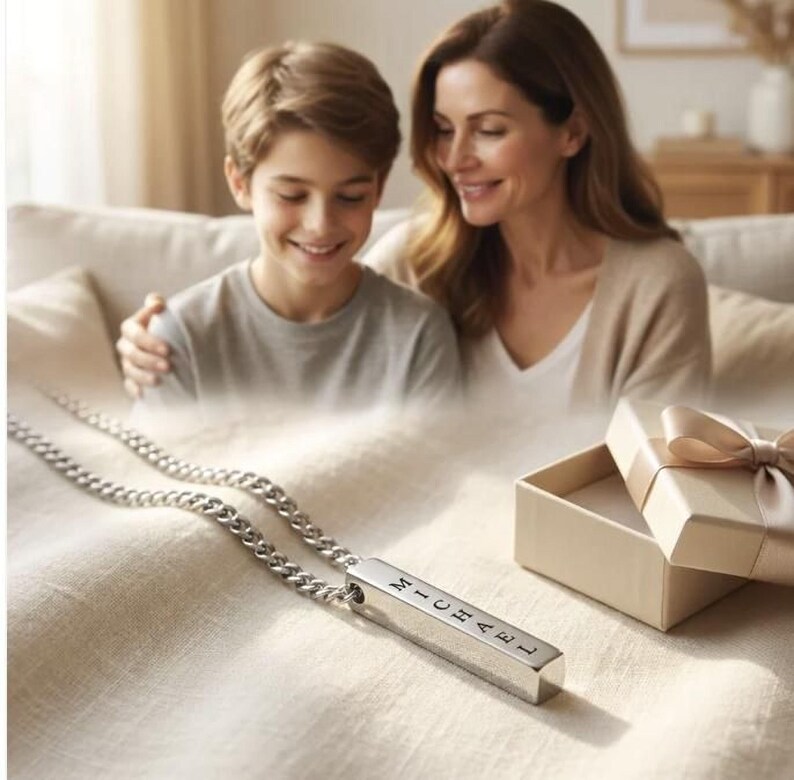 A mother and son are sitting together on a couch, smiling and looking at a necklace with a pendant that has the name "Michael" engraved on it.