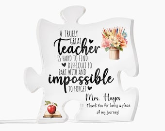 May include: A white acrylic puzzle piece shaped like a teacher appreciation gift. The puzzle piece has a black and white graphic with the text "A truly great teacher is hard to find, difficult to part with and impossible to forget."  The graphic also includes a red apple on a book, a vase of flowers, and pencils. The text "Mrs. Hayes Thank You for being a piece of my journey!" is also included.