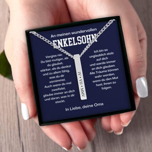 a person's hand holding a necklace with a black box containing a message in German.