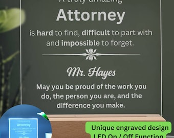 May include: A personalized Attorney gift. The clear acrylic plaque features engraved text: "A truly amazing Attorney...impossible to forget." It includes the name "Mr. Hayes" and a motivational message. The plaque sits on a wooden base with LED lighting.