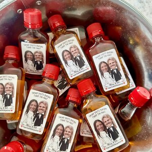 May include: A bucket filled with small glass bottles with red caps. Each bottle has a personalized label with a cartoon image of a couple and the text "Taking Shots Before Tying The Knot" with the couple's names and a hashtag.