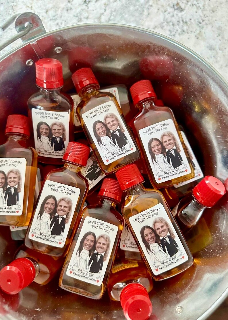 May include: A bucket filled with small glass bottles with red caps. Each bottle has a personalized label with a cartoon image of a couple and the text "Taking Shots Before Tying The Knot" with the couple's names and a hashtag.