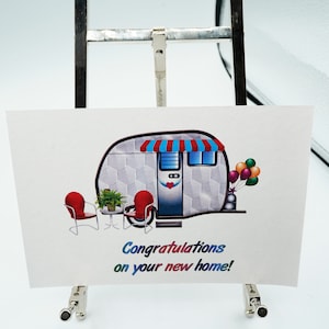 May include: A white and gray camper with a red and blue awning, two red chairs, a small table with a green plant, and a bunch of colorful balloons. The text "Congratulations on your new home!" is printed below the camper.
