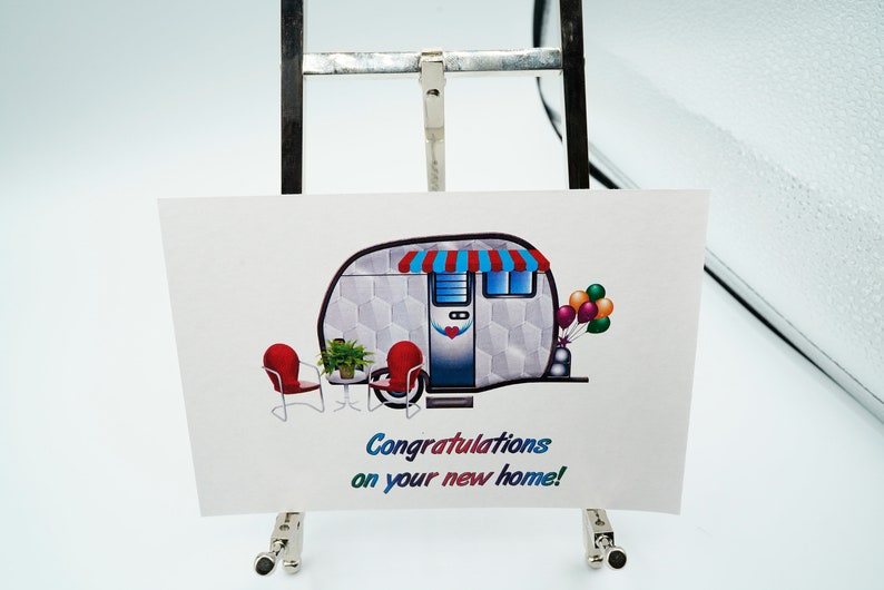 May include: A white and gray camper with a red and blue awning, two red chairs, a small table with a green plant, and a bunch of colorful balloons. The text "Congratulations on your new home!" is printed below the camper.