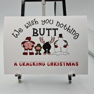 Peut inclure: Une carte blanche avec le texte "We wish you nothing BUTT" et "A Cracking Christmas" en rouge et noir. La carte pr&eacute;sente des fesses de dessins anim&eacute;s habill&eacute;es en P&egrave;re No&euml;l, elfe, renne et bonhomme de neige.
