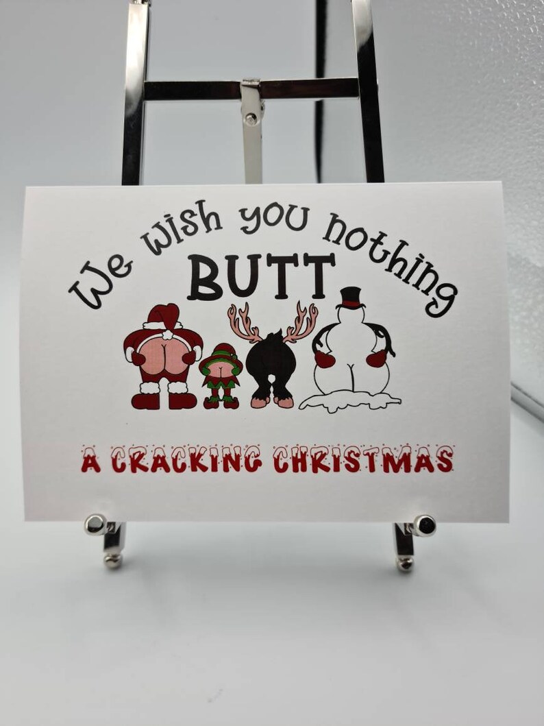 Peut inclure: Une carte blanche avec le texte "We wish you nothing BUTT" et "A Cracking Christmas" en rouge et noir. La carte pr&eacute;sente des fesses de dessins anim&eacute;s habill&eacute;es en P&egrave;re No&euml;l, elfe, renne et bonhomme de neige.
