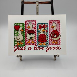 May include: A white card with the text "Just a love goose" in red script. The card features four embroidered geese, each in a different pose and holding a heart, flowers, a bow, and a mug. The geese are set against colorful, patterned backgrounds.