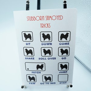 May include: A white card with the text "Stubborn Samoyed Tricks" printed in red and black. The card has a grid of nine squares, each containing a black silhouette of a Samoyed dog and a word describing a dog trick, such as "sit", "down", "come", "shake", "roll over", "go", "fetch", "jump", "paw", "go to bed", and "stay".