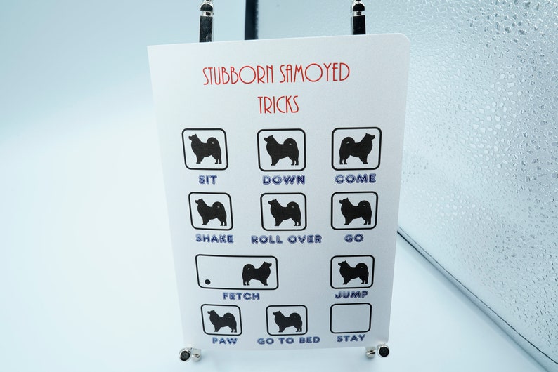 May include: A white card with the text "Stubborn Samoyed Tricks" printed in red and black. The card has a grid of nine squares, each containing a black silhouette of a Samoyed dog and a word describing a dog trick, such as "sit", "down", "come", "shake", "roll over", "go", "fetch", "jump", "paw", "go to bed", and "stay".