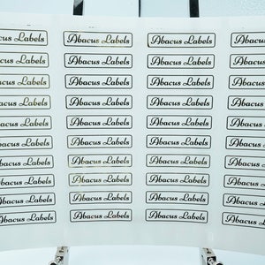 Może przedstawiać: Arkusz białego papieru z 36 złotymi etykietami, na których nadrukowano tekst "Abacus Labels" czarną czcionką kursywną.
