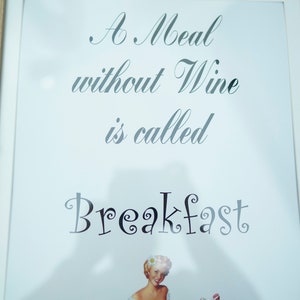 Op de afbeelding: Een ingelijst afdruk met een witte achtergrond en zwarte tekst die "A Meal without Wine is called Breakfast" luidt. Rechtsonder staat een pin-up-illustratie van een vrouw in een rode rok en een wit topje die een fles wijn en een picknickmand vasthoudt.