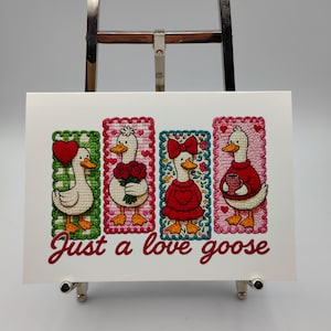 May include: A framed artwork featuring four embroidered geese, each in a different pose and outfit, set against colorful backgrounds. The phrase "Just a love goose" is written in cursive below the geese. The artwork is displayed on a silver easel.