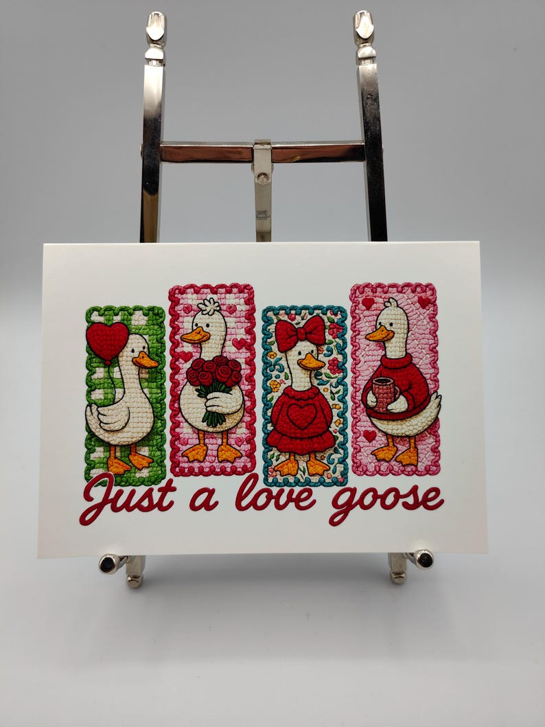 May include: A framed artwork featuring four embroidered geese, each in a different pose and outfit, set against colorful backgrounds. The phrase "Just a love goose" is written in cursive below the geese. The artwork is displayed on a silver easel.