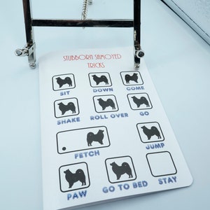 May include: A white card with black silhouettes of Samoyed dogs and the text "Stubborn Samoyed Tricks" at the top. The card lists various dog tricks such as "Sit", "Down", "Come", "Shake", "Roll Over", "Go", "Fetch", "Jump", "Paw", "Go to Bed", and "Stay".