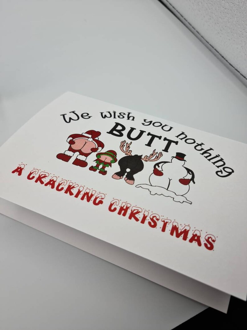 Peut inclure: Carte de No&euml;l blanche avec le texte &laquo;&nbsp;We wish you nothing BUTT a Cracking Christmas&nbsp;&raquo;. La carte pr&eacute;sente des illustrations de dessins anim&eacute;s du P&egrave;re No&euml;l, d'un elfe, d'un renne et d'un bonhomme de neige, chacun avec son post&eacute;rieur expos&eacute;.
