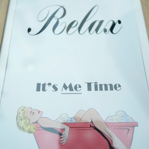 Op de afbeelding: Ingelijste print met het woord "Relax" in elegant schrift boven de zin "It's Me Time". De afbeelding toont een pop-art-stijl illustratie van een vrouw die ontspant in een rode badkuip vol bubbels. De lijst is lichtbruin.