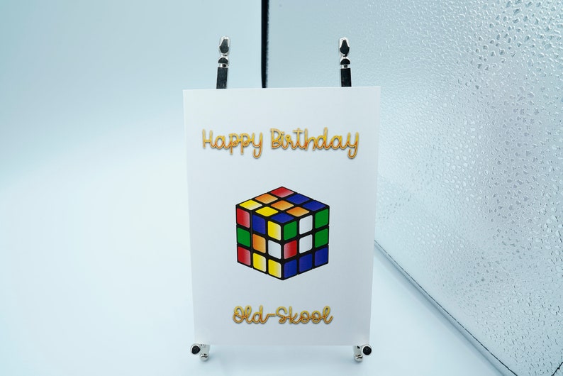 May include: A white card with the text "Happy Birthday" in gold lettering. A colorful Rubik's Cube is below the text, and the text "Old-School" is below the cube in gold lettering.
