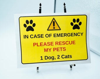 Pode incluir: Uma placa amarela com pegadas pretas e o texto "IN CASE OF EMERGENCY PLEASE RESCUE MY PETS 1 Dog, 2 Cats". Um símbolo de ponto de exclamação preto está em um triângulo amarelo. A placa é presa por dois clipes de metal.