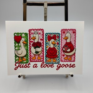 May include: A white card featuring four embroidered geese in rectangular frames. Each goose has a different accessory: a heart, flowers, a bow, and a mug. The text "Just a love goose" is written in red script below the geese.