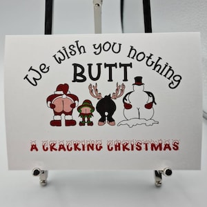 Peut inclure: Une carte blanche avec le texte "We wish you nothing BUTT" et "A Cracking Christmas" en rouge et noir. La carte pr&eacute;sente des images de dessins anim&eacute;s d'un P&egrave;re No&euml;l, d'un elfe, d'un orignal et d'un bonhomme de neige, chacun avec son post&eacute;rieur expos&eacute;.