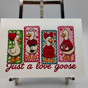 May include: A white card featuring four embroidered goose illustrations, each within a decorative frame. The geese hold a heart balloon, flowers, a bow, and a mug. The text "Just a love goose" is written in red script.
