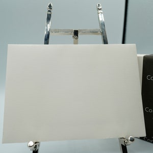 May include: A white textured paper sheet is displayed on a silver easel. The paper is rectangular and has a subtle horizontal lined pattern. A black and white box with the text "Conqueror" is visible.