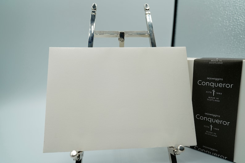 May include: A white textured paper sheet is displayed on a silver easel. The paper is rectangular and has a subtle horizontal lined pattern. A black and white box with the text "Conqueror" is visible.