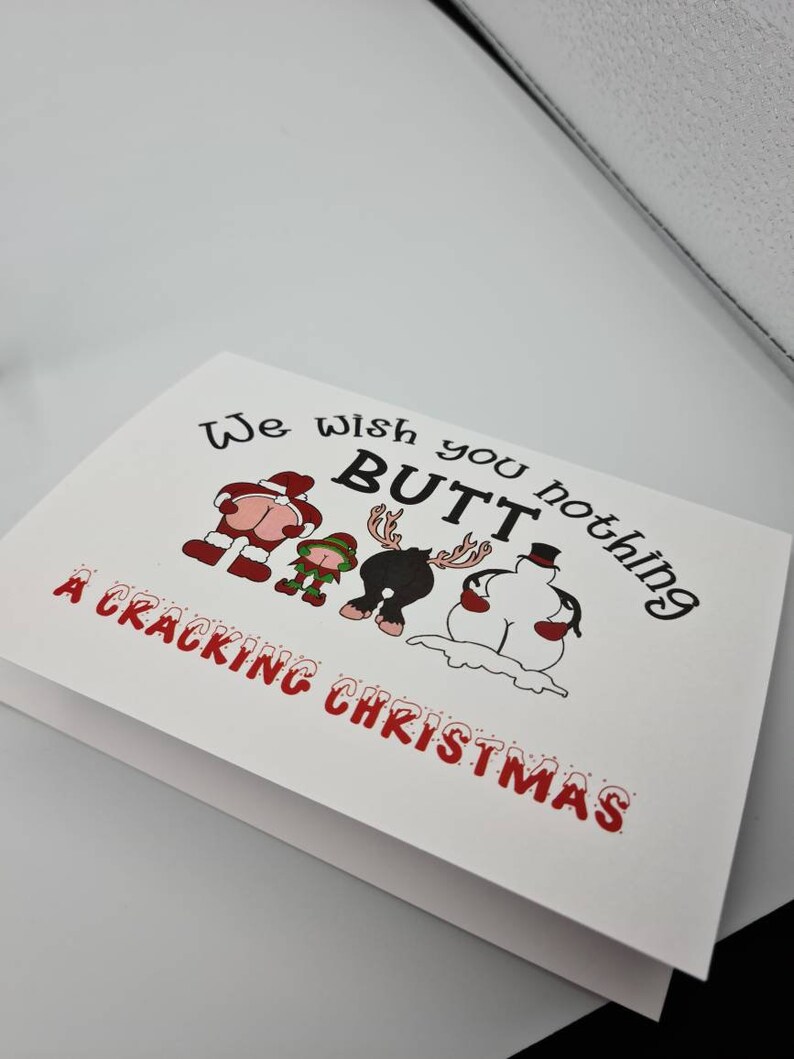 Peut inclure: Carte de No&euml;l blanche avec le texte "We wish you nothing BUTT a Cracking Christmas." La carte pr&eacute;sente des images de dessins anim&eacute;s du P&egrave;re No&euml;l, d'un elfe, d'un renne et d'un bonhomme de neige, chacun avec son post&eacute;rieur expos&eacute;.