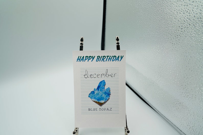May include: A white card with blue and green text that reads "HAPPY BIRTHDAY December BLUE TOPAZ". A blue topaz crystal is pictured on the card.