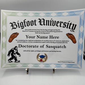 Op de afbeelding: Een novelty diploma van Bigfoot University. Het certificaat heeft een groene en blauwe rand en toont de naam van de universiteit in gotische letters. De tekst luidt "Doctorate of Sasquatch" en bevat een ruimte voor een naam.