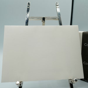 May include: A white card with a textured surface is displayed on a silver easel. The card is blank. The card is packaged in a black box with the text "Arjowiggins Conqueror Est. 1888 Made in Scotland" printed on it.