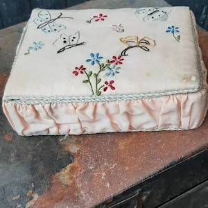 May include: A small, rectangular, pink and white embroidered box with a ruffled edge. The embroidery features butterflies, flowers, and leaves in shades of red, blue, green, and yellow.