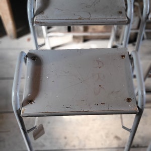 May include: Two gray metal stools with a white painted surface. The top stool has the number 485 written on it.