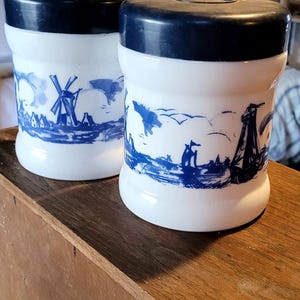 May include: Two white ceramic canisters with blue and white windmill and boat designs. The canisters have blue lids.