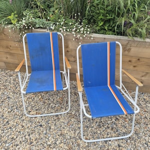 May include: Two blue folding chairs with white frames and wooden armrests. Each chair has a vertical orange and white stripe down the centre of the blue seat back. The chairs are on a bed of small stones.