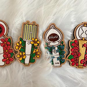 May include: Four wooden pins with floral designs. The pins feature a variety of shapes, including a compass, a microphone, a light bulb with the word "xikers", and an hourglass. Each pin is decorated with flowers in red, yellow, and blue.