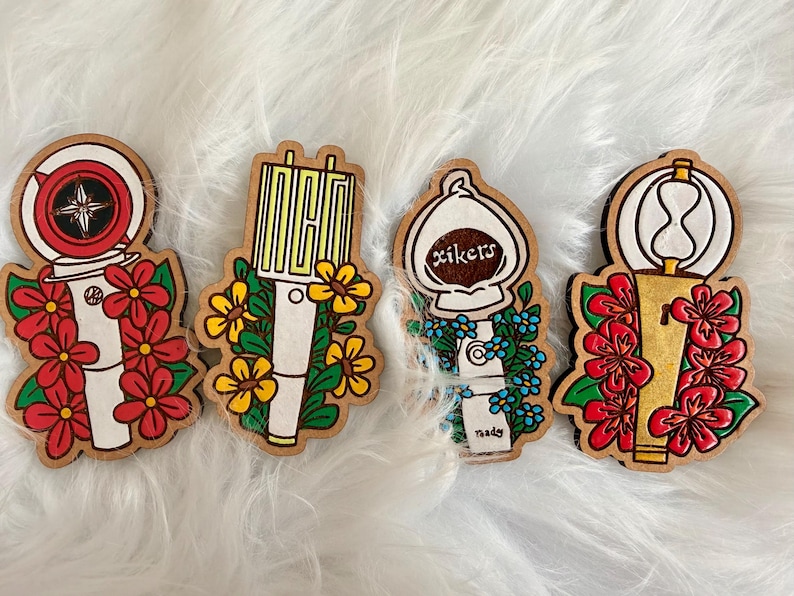 May include: Four wooden pins with floral designs. The pins feature a variety of shapes, including a compass, a microphone, a light bulb with the word "xikers", and an hourglass. Each pin is decorated with flowers in red, yellow, and blue.
