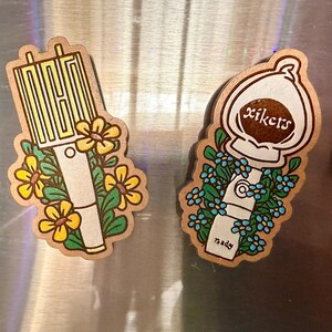 May include: Two wooden stickers with floral designs. One features the word "NCT" in yellow, surrounded by yellow flowers. The other has the word "xikers" and blue flowers. Both have a white and beige color scheme.