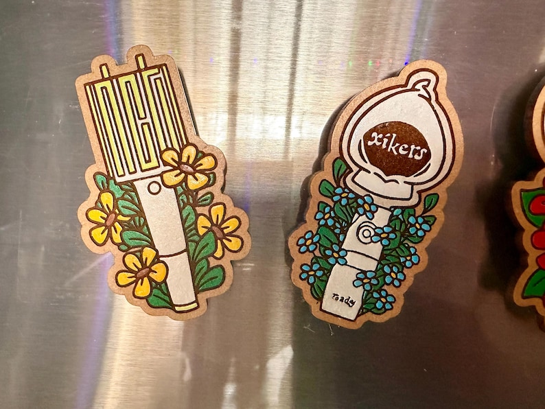 May include: Two wooden stickers with floral designs. One features the word "NCT" in yellow, surrounded by yellow flowers. The other has the word "xikers" and blue flowers. Both have a white and beige color scheme.