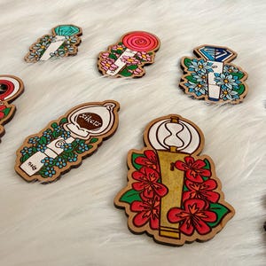 May include: A collection of wooden pins with various designs. Some pins feature floral arrangements, while others display geometric shapes and text. The pins are painted with bright colors, including red, blue, and green, and have a natural wood border.