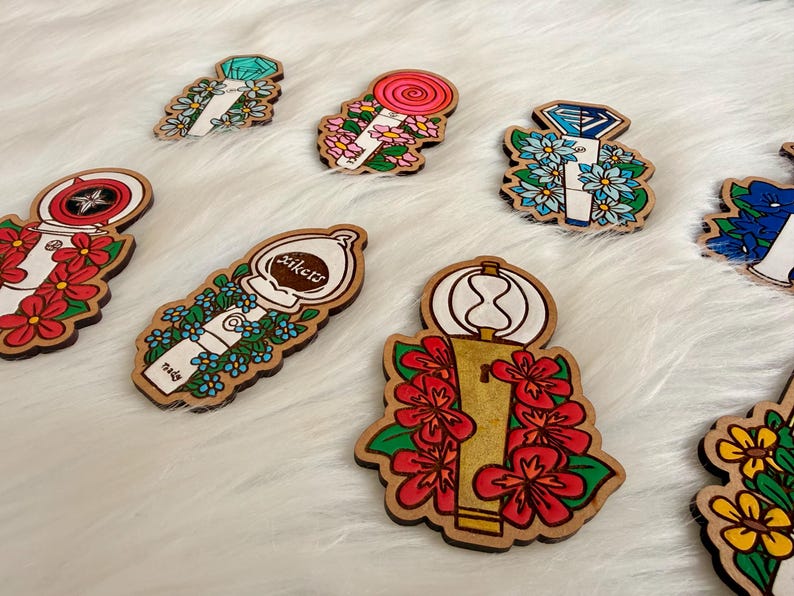 May include: A collection of wooden pins with various designs. Some pins feature floral arrangements, while others display geometric shapes and text. The pins are painted with bright colors, including red, blue, and green, and have a natural wood border.