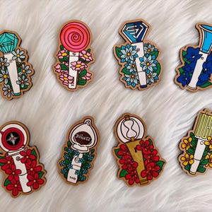 May include: A collection of eight wooden fan light stick pins, each with a unique design. The pins feature various shapes and colors, including a lollipop, a diamond, and a horn, all adorned with floral accents. The pins are set against a white, fluffy background.