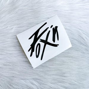 Puede incluir: Una pegatina rectangular blanca con un dise&ntilde;o gr&aacute;fico negro audaz y de estilo dibujado a mano. La obra consiste en l&iacute;neas y trazos negros gruesos, expresivos y entrecruzados que forman una forma abstracta y estilizada sobre el fondo blanco.