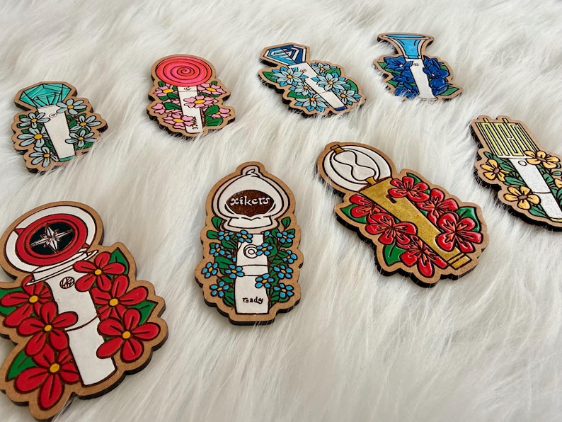 May include: Wooden pins with various designs, including a lollipop, a diamond, and a compass, each surrounded by floral accents. The pins are painted in bright colors and have a natural wood border.