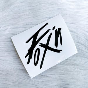 Puede incluir: Una pegatina rectangular blanca con un dise&ntilde;o gr&aacute;fico negro audaz que se asemeja a una firma estilizada dibujada a mano o a letras abstractas. El dise&ntilde;o consiste en trazos de tinta negra afilados y angulares en un patr&oacute;n din&aacute;mico.