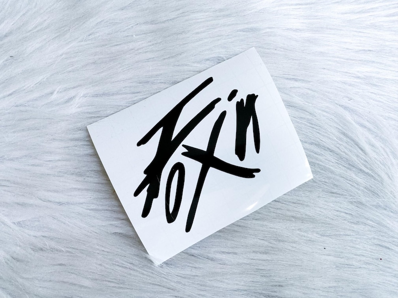 Puede incluir: Una pegatina rectangular blanca con un dise&ntilde;o gr&aacute;fico negro audaz que se asemeja a una firma estilizada dibujada a mano o a letras abstractas. El dise&ntilde;o consiste en trazos de tinta negra afilados y angulares en un patr&oacute;n din&aacute;mico.