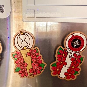 May include: Wooden magnets with a floral design. One magnet features a hourglass shape, and the other has a compass design. Both magnets are decorated with red flowers and green leaves.